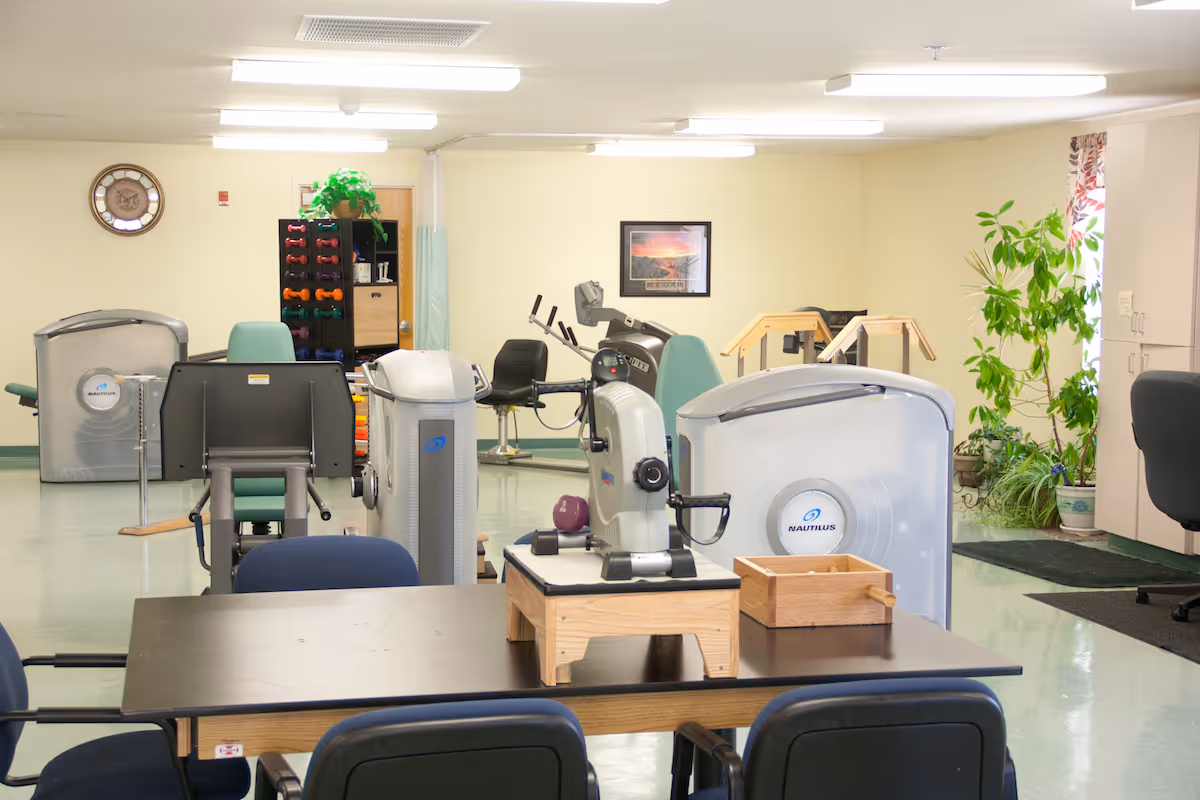 Rehabilitation exercise room with fitness machines, hand weights, chairs, and a table.