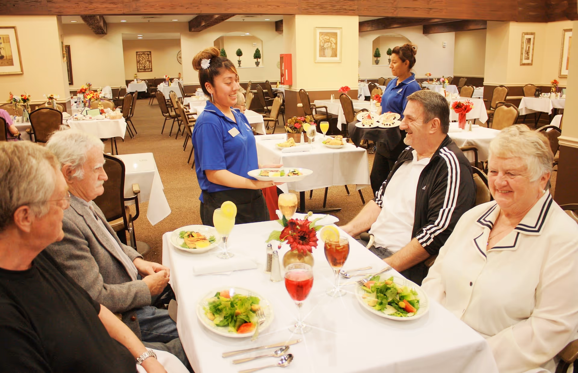 A group of elderly people sitting around a table in a dining room at a retirement community, being served by two staff members in blue uniforms. The table is set with plates of salad and drinks, and the room has multiple tables with white tablecloths and floral centerpieces.