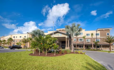 Exterior view of a multi-story senior living facility building with a well-maintained lawn and palm trees under a partly cloudy blue sky.