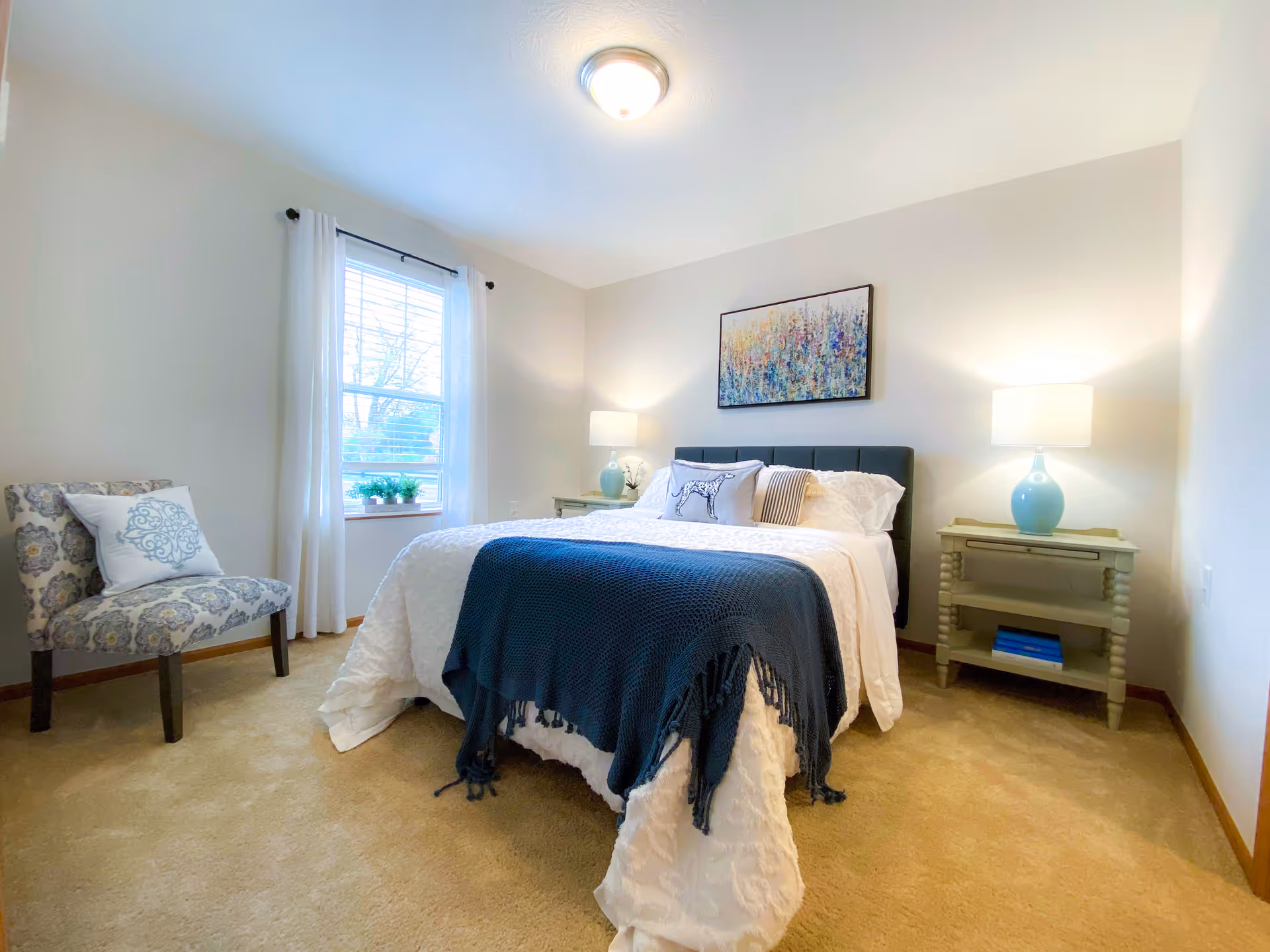 A bright bedroom with a bed covered in white bedding and a dark blue throw blanket. There are two nightstands with blue lamps on either side of the bed, a colorful abstract painting above the bed, a window with white curtains, and a patterned chair with a decorative pillow near the window.