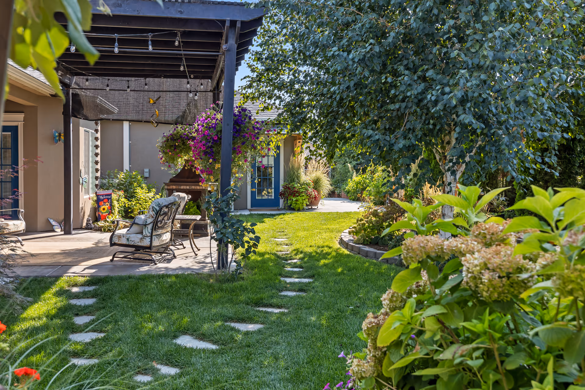 Sunny backyard garden patio with a pergola, cushioned seating, hanging flower baskets and a stepping-stone lawn path.