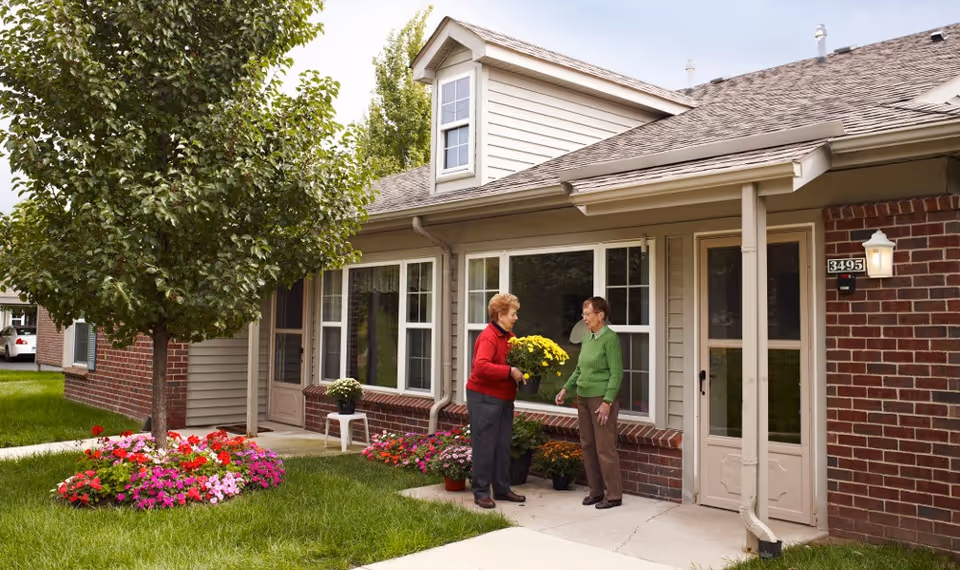 Two older women standing in front of a single-story brick-and-siding senior living unit, one holding a pot of yellow flowers beside a tree and flowerbeds.