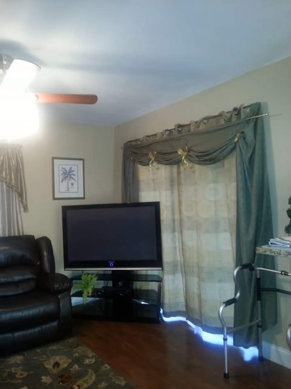 Living room with a flat-screen TV on a glass stand, a leather recliner, decorative curtains and a walker by the sliding door.