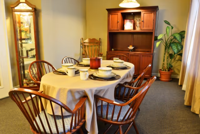 A dining room with a wooden oval table covered with a beige tablecloth, set with six place settings including bowls, plates, and mugs. Surrounding the table are six wooden chairs with cushions. In the background, there is a wooden cabinet with glass doors and shelves, a tall wooden rocking chair, a potted plant, and beige curtains covering a window.