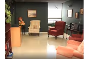 A cozy living room with green walls, featuring a beige armchair, a red armchair, and a peach-colored sofa. There is a small wooden table with a vintage radio and a lamp, a bookshelf on the left, and framed pictures on the walls. A window with blinds is visible in the background.