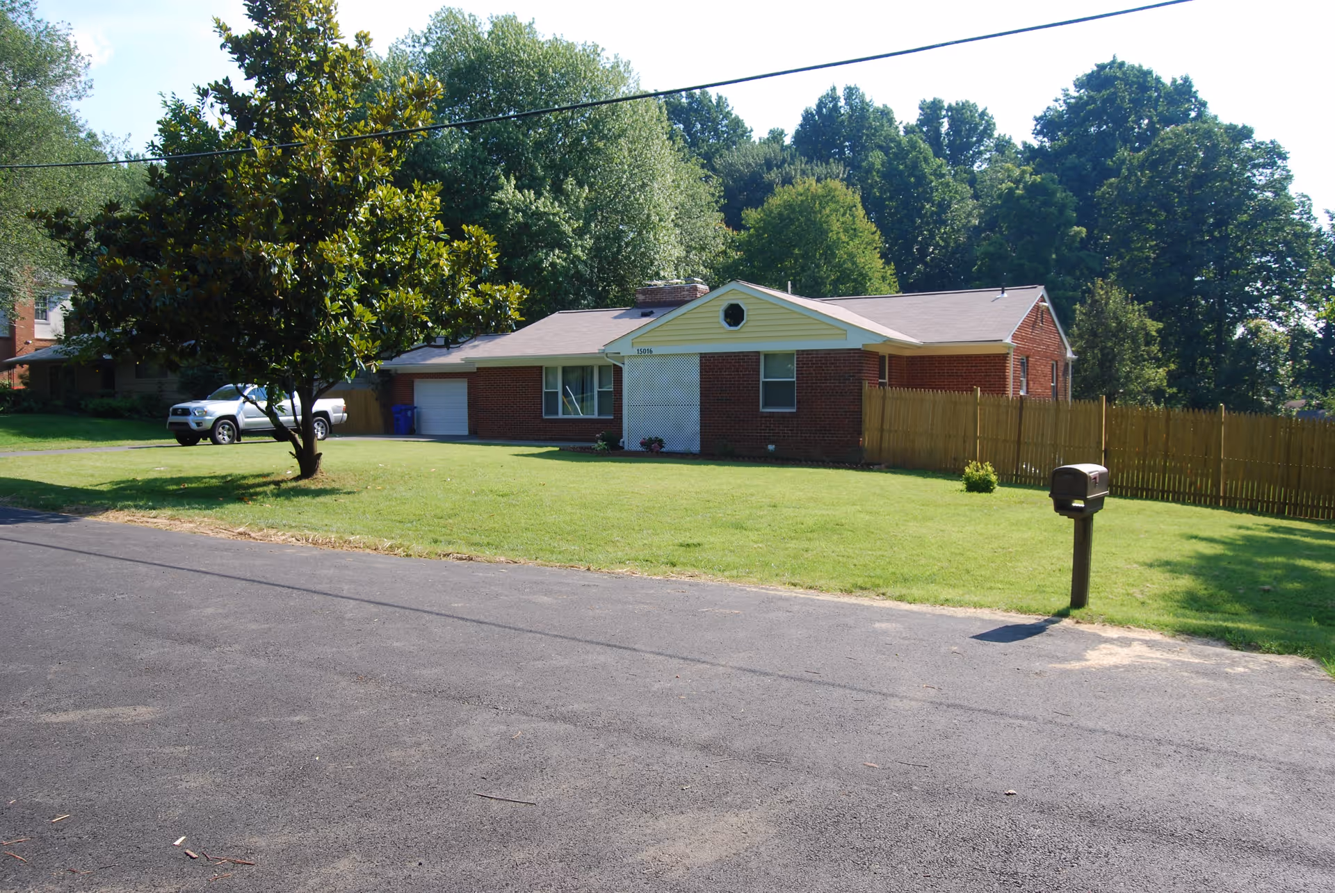 Single-story brick house with a front lawn, a tree, a mailbox, and a driveway with a parked white vehicle. The house has a fenced backyard and is surrounded by trees.