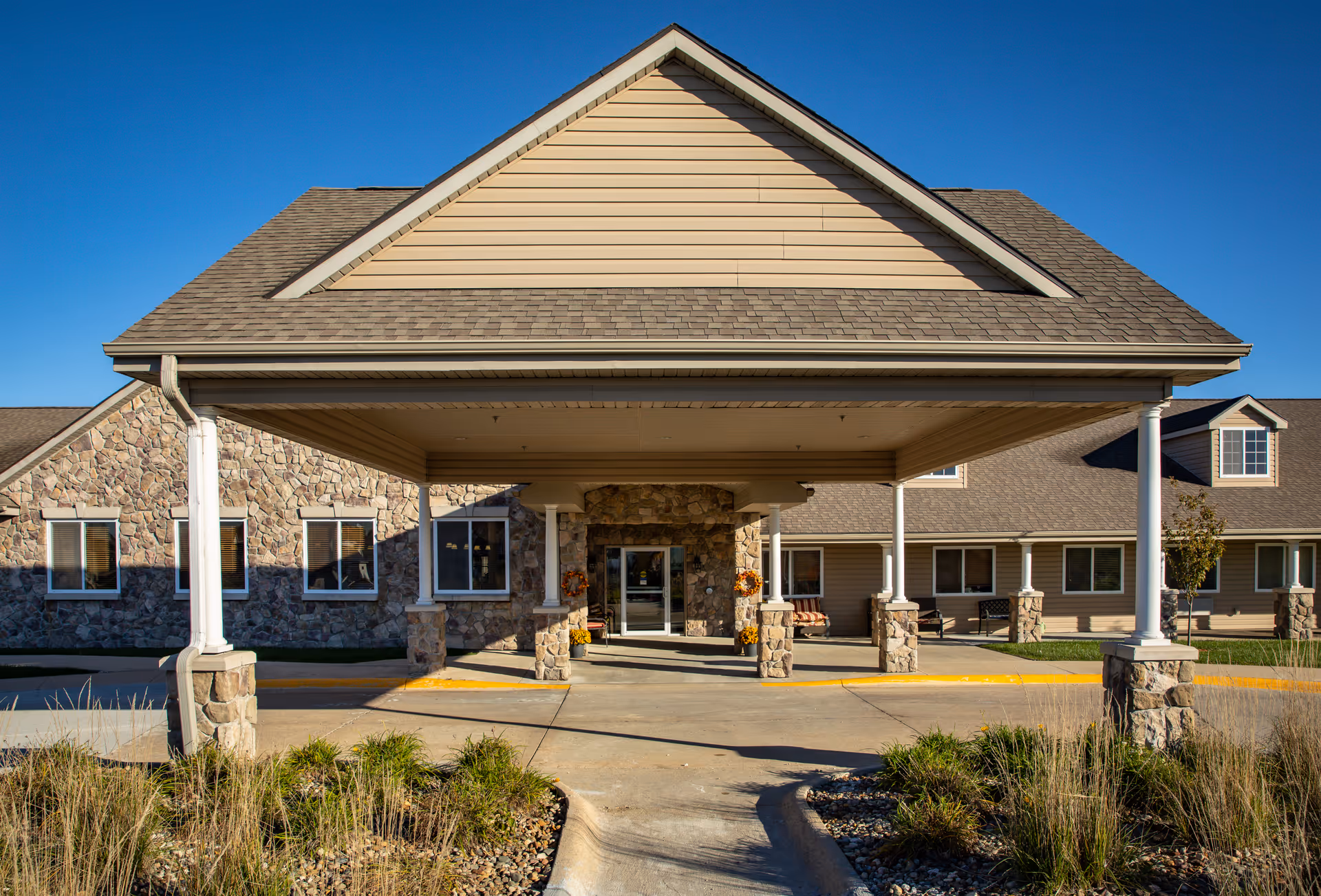 Front exterior view of Homestead Assisted Living & Memory Care of Creston building with a covered entrance supported by stone pillars, stone and siding facade, and a clear blue sky.