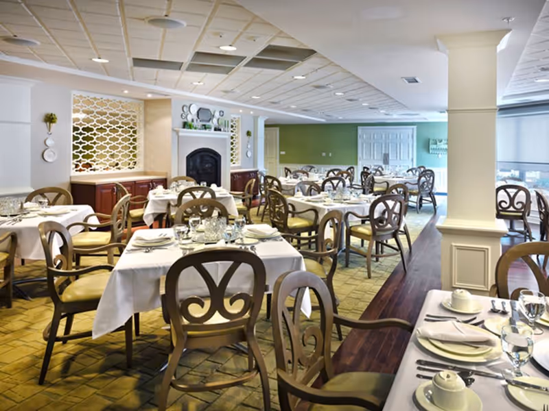 A bright and elegant dining room with multiple tables covered in white tablecloths, set with plates, glasses, and silverware. The room features wooden chairs with decorative backs, a fireplace with a mantel adorned with plates and greenery, and a mix of tiled and wooden flooring. The walls are painted in light colors with some green accents, and there are large windows letting in natural light.