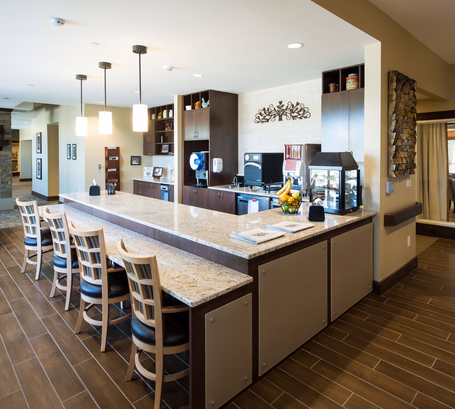A modern kitchen area with a long granite countertop and four wooden chairs with black cushions lined up along the counter. The kitchen features dark wood cabinets, a coffee machine, a fruit basket with bananas and oranges, and decorative items on shelves. Pendant lights hang from the ceiling above the counter, and the floor is covered with brown tiles.