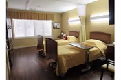 A well-lit bedroom in a healthcare facility featuring two hospital-style beds with yellow bedding, a wooden floor, a large window with blinds and a valance, a chair, and a small table between the beds.