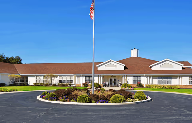 Front exterior view of Kessler Estates Senior Living building with a circular driveway, landscaped garden in the center with bushes and flowers, and an American flag on a tall flagpole in front of the entrance.