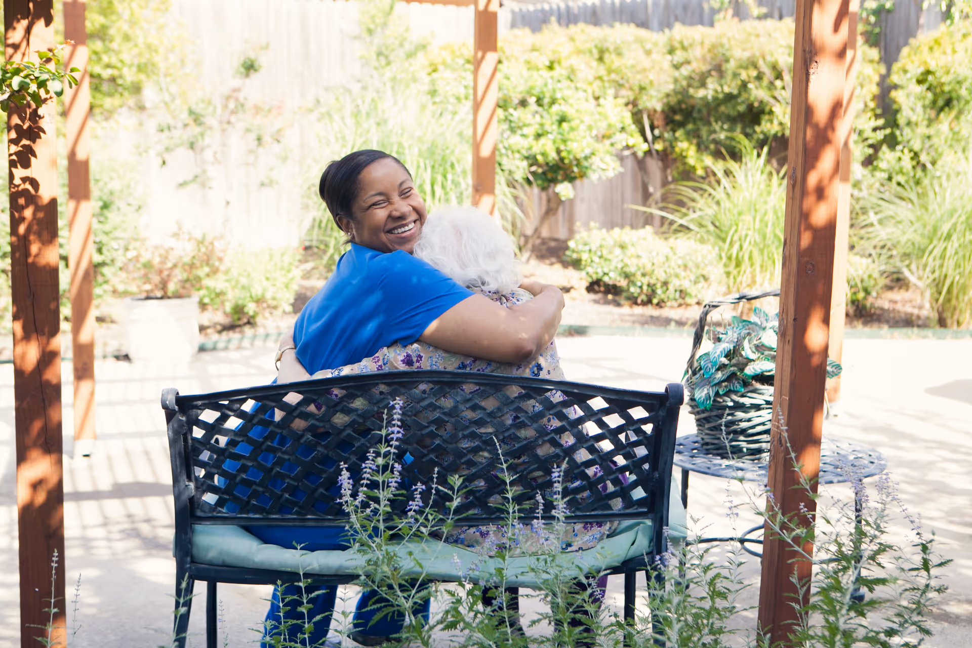 A smiling caregiver in blue scrubs hugs an elderly woman with white hair while sitting together on a black metal bench in a sunny garden patio area with plants and wooden posts.