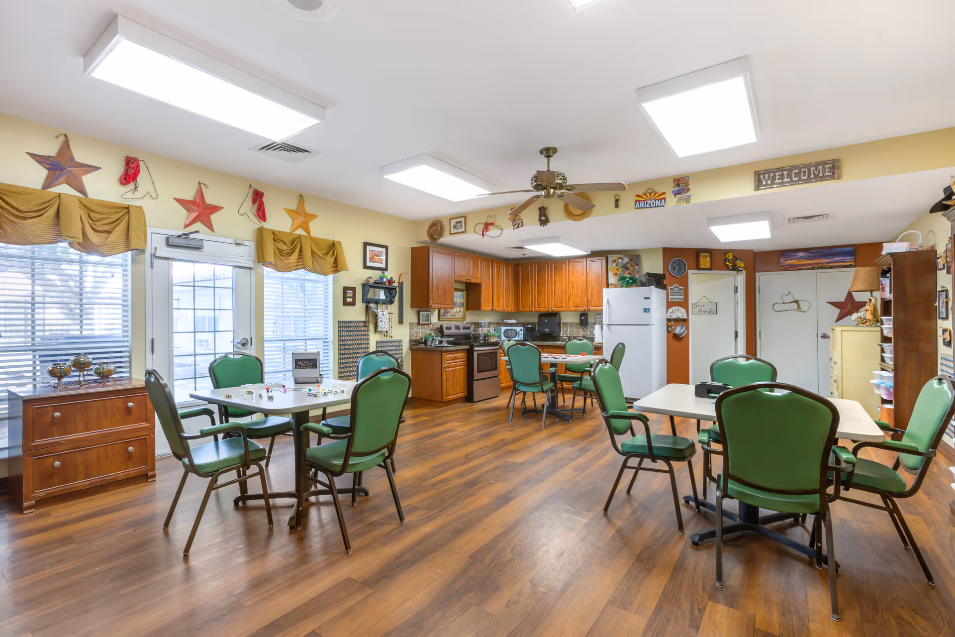 A bright and spacious common area with a kitchen and dining space. The room features wooden flooring, green cushioned chairs around tables, wooden kitchen cabinets, a white refrigerator, and various decorative items on the walls including stars, a welcome sign, and Arizona-themed decor. Large windows with yellow valances allow natural light to enter the room.