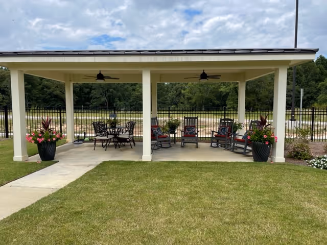 Outdoor covered seating area with a concrete floor, featuring a table with four metal chairs on the left and four rocking chairs with cushions on the right. Two ceiling fans are mounted on the roof of the structure. Large potted plants with flowers are placed at the front corners of the structure. The area is surrounded by a black metal fence with trees and a partly cloudy sky in the background.