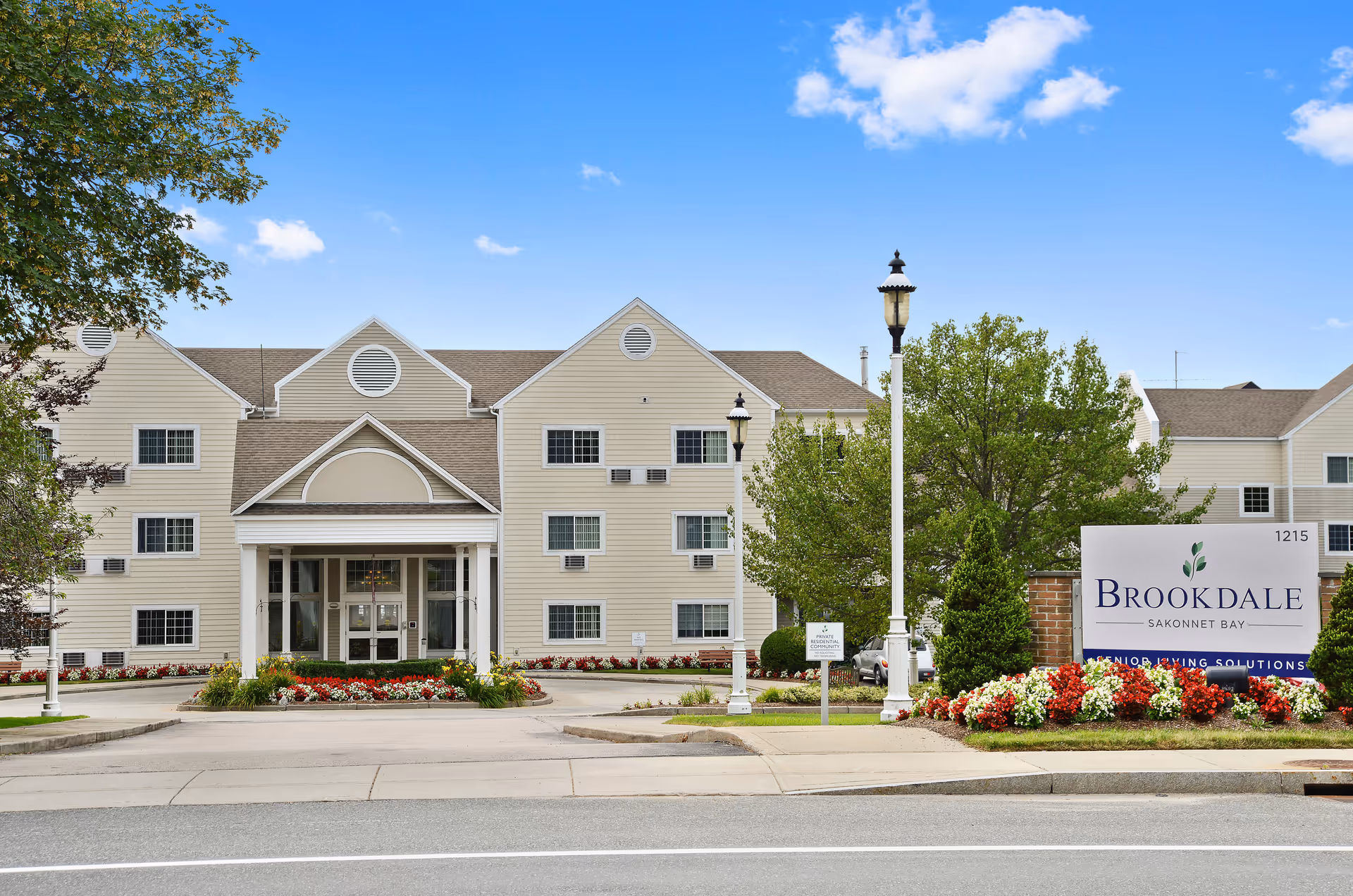Front exterior of the Brookdale Sakonnet Bay senior living building with a covered entrance, lamp posts, and landscaped flowerbeds.