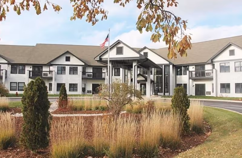 Exterior view of a two-story senior living facility building with white walls and a gray roof, surrounded by landscaped bushes, tall grasses, and a circular driveway. An American flag is flying on a flagpole near the entrance, and tree branches with autumn leaves frame the top of the image.