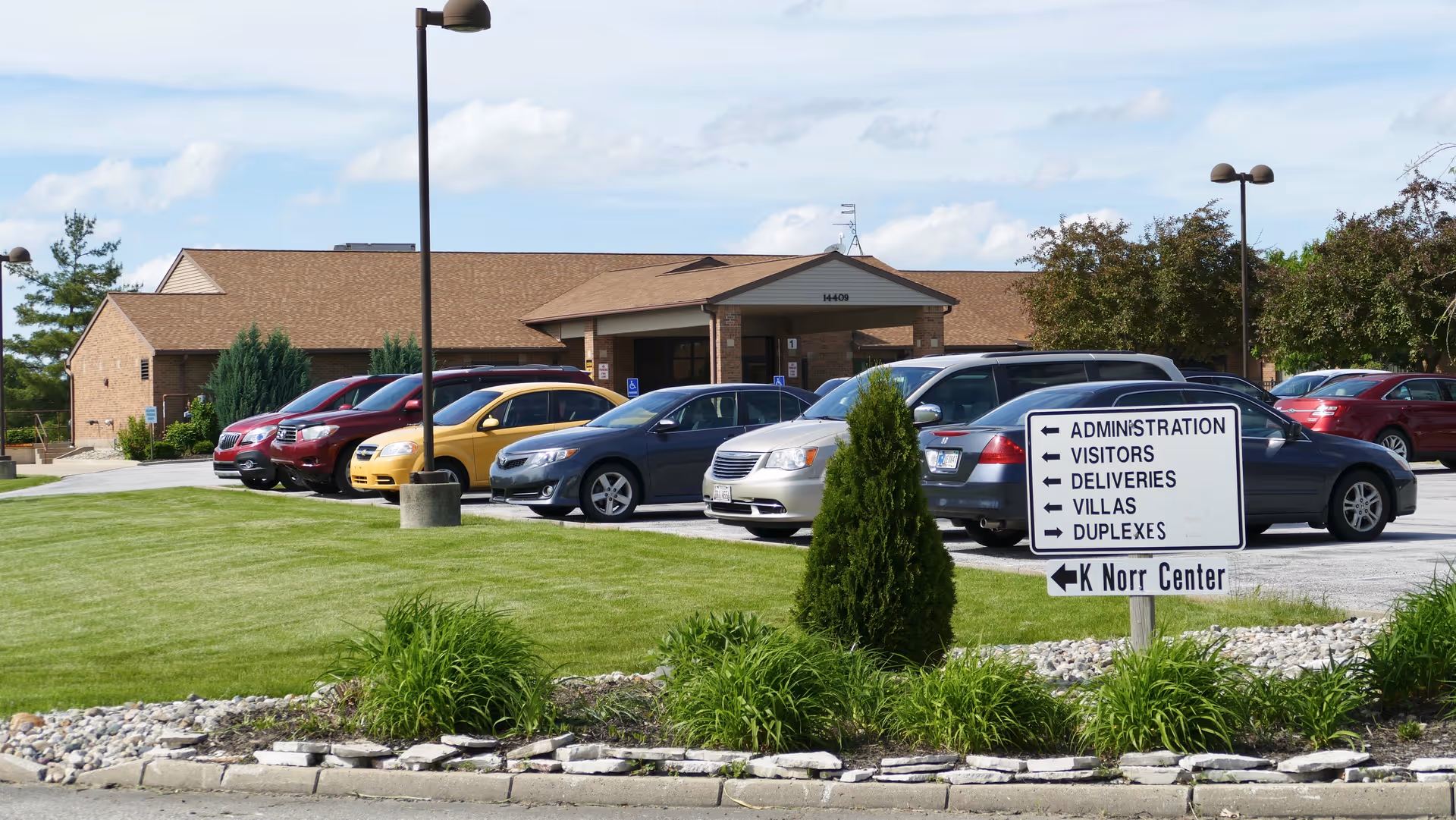Parking lot with several cars parked in front of a single-story brick building with a brown roof. A sign in the foreground directs to administration, visitors, deliveries, villas, duplexes, and K Norr Center. The area is landscaped with grass, small bushes, and rocks.
