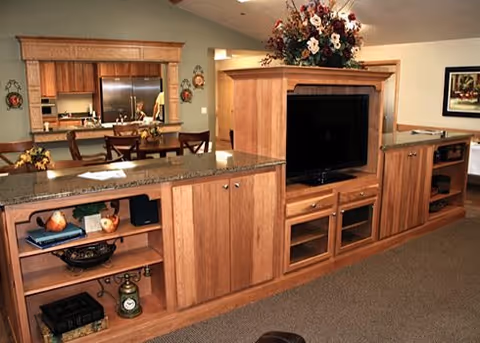 Open communal living area featuring a large wooden media cabinet with a flat-screen TV, granite-topped counters, and a view into a dining area and kitchen.