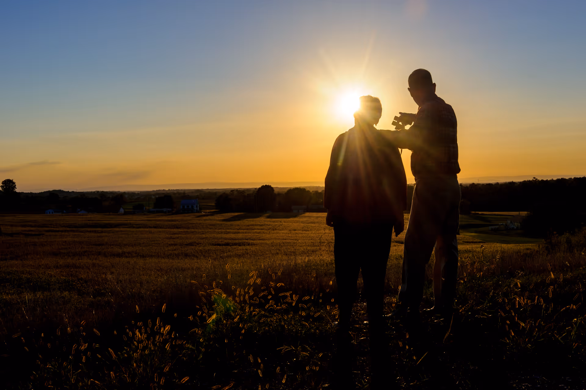 Silhouettes of two people standing in a field during sunset, with one person pointing towards the horizon and the sun shining brightly behind them.
