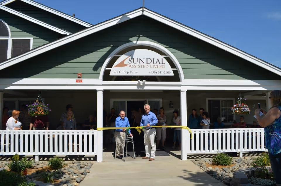 Front exterior view of Sundial Assisted Living facility with people gathered for a ribbon-cutting ceremony. Two men stand in the center holding a yellow ribbon, one using a walker. The building has green siding, white trim, and hanging flower baskets on the porch.