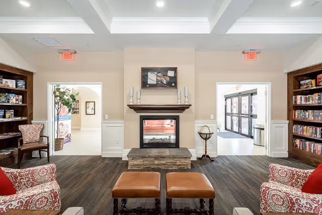 A cozy living room area in Fellowship Square Surprise featuring a central fireplace with a stone base and a wooden mantle holding four candles. Above the fireplace is a framed picture. On either side of the fireplace are doorways leading to other parts of the facility. The room has built-in wooden bookshelves filled with books and games, patterned armchairs with red cushions, and two brown leather ottomans in front of the fireplace. The ceiling has recessed lighting and a tray design.