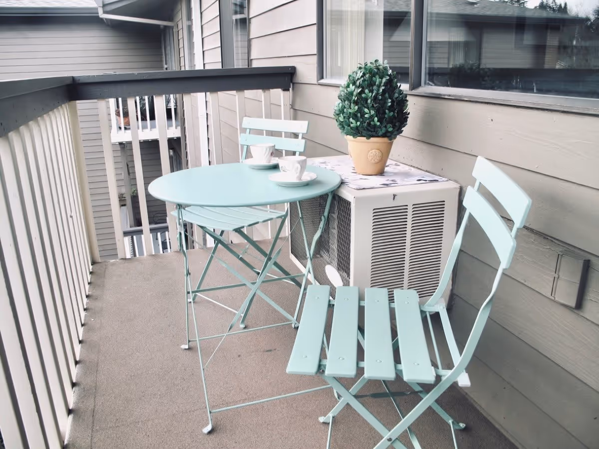 Small outdoor balcony with a light blue metal table and two matching chairs. On the table are two white cups and saucers. A potted green plant sits on top of an air conditioning unit against the beige siding of the building.