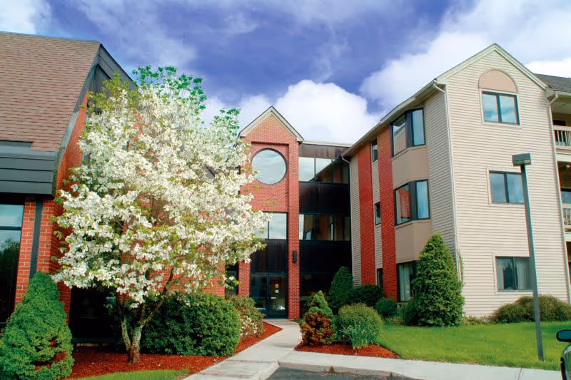 Exterior view of a senior living facility named East Hill, featuring a multi-story building with a combination of brick and beige siding, surrounded by well-maintained landscaping including a blooming white-flowered tree and green shrubs under a partly cloudy sky.