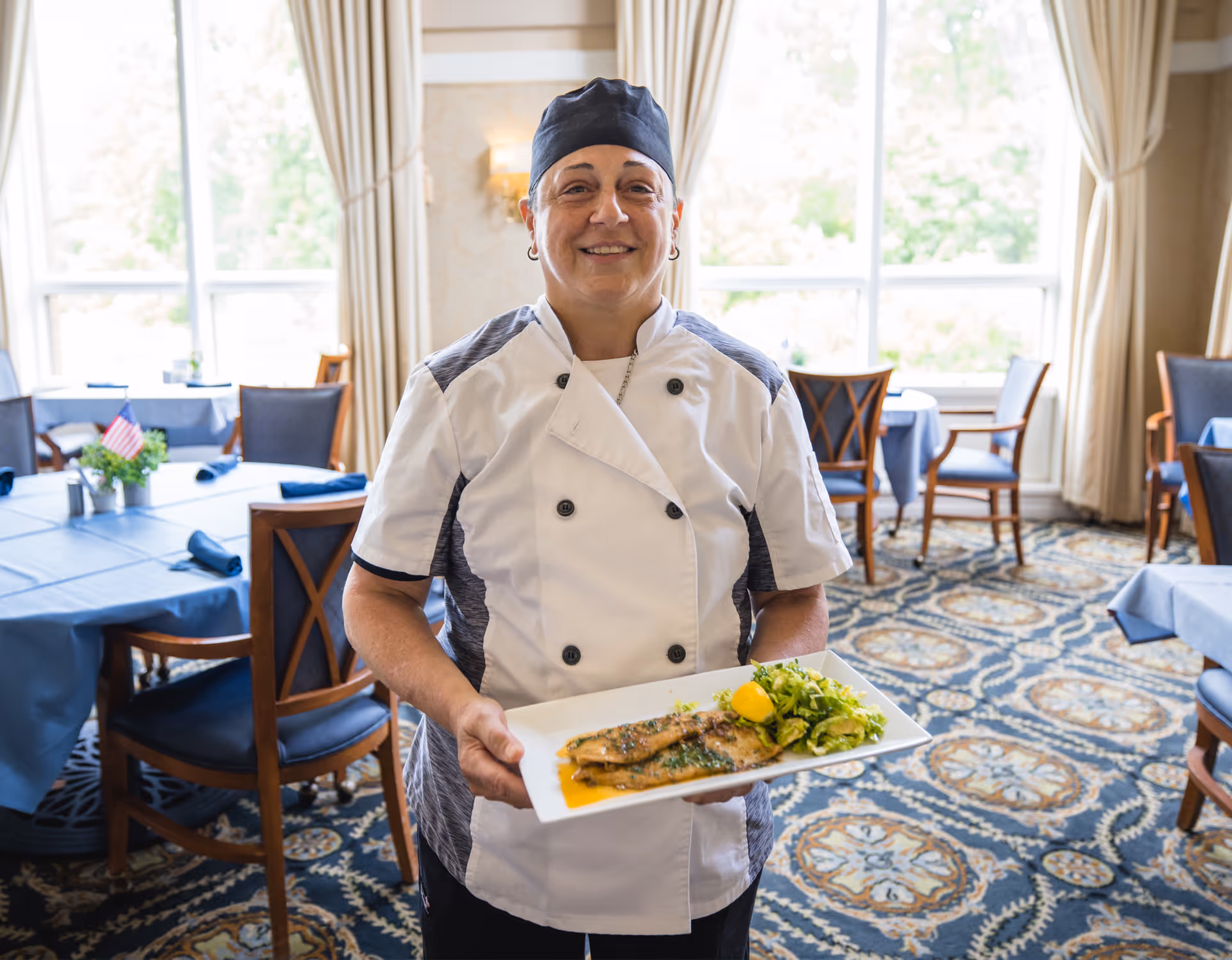 A chef wearing a white and gray uniform and black cap is smiling and holding a rectangular white plate with a cooked fish fillet garnished with herbs, a lemon wedge, and a side of green salad. The background shows a dining room with tables covered in blue tablecloths, wooden chairs, large windows with beige curtains, and a patterned carpet.