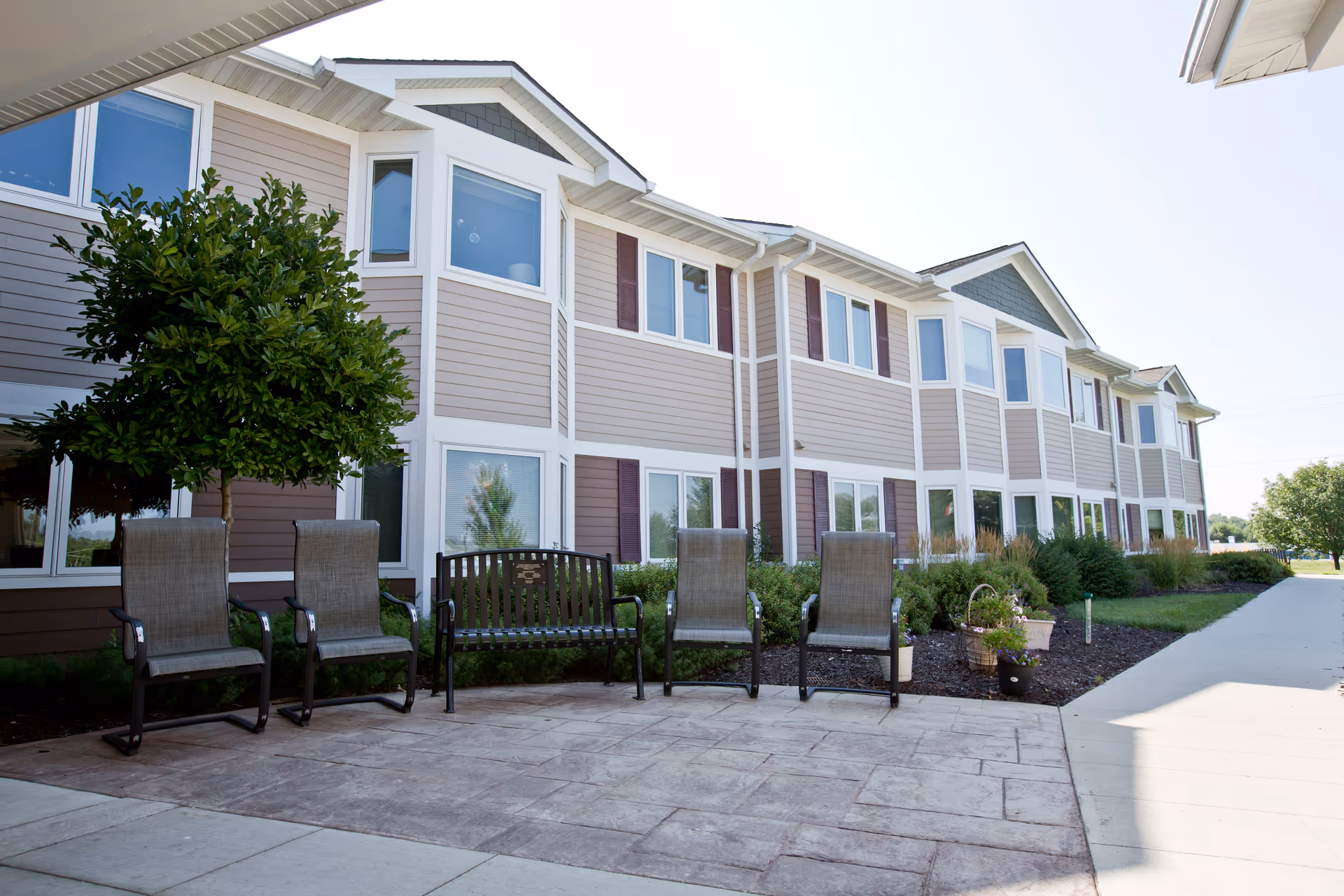 Outdoor patio area with several chairs and a bench in front of a two-story building with multiple windows and beige siding. There are small trees and landscaped bushes along the building.