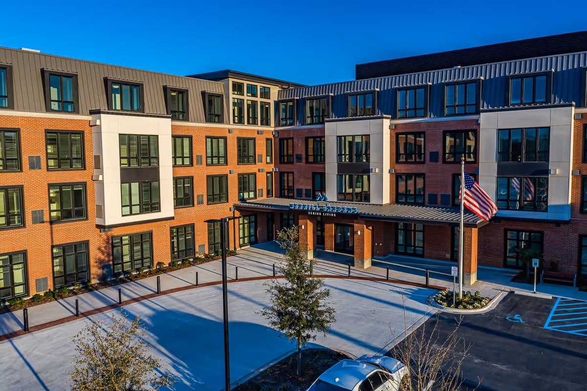 Exterior view of Merrill Gardens at Columbia senior living facility showing a modern multi-story brick building with large windows, an entrance canopy, an American flag on a flagpole, and a parking area with a handicap parking space.