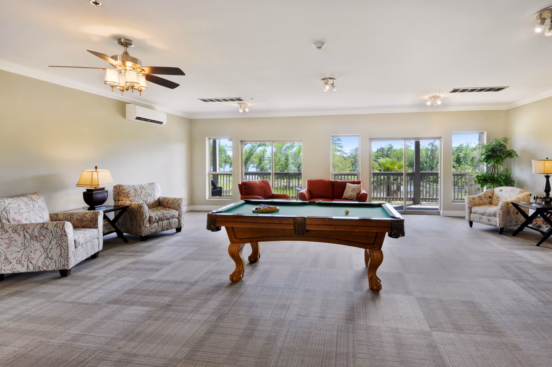 Spacious bright common room featuring a pool table in the center, sofas and armchairs, and large windows with a view of trees.