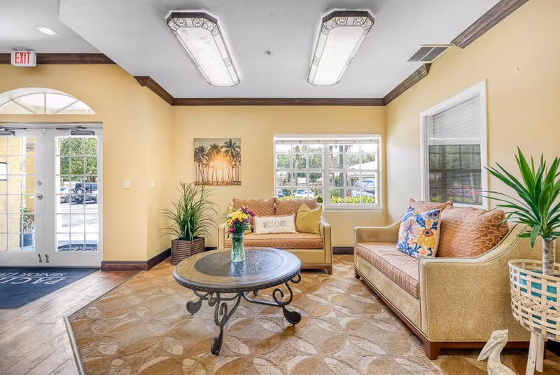 A bright and welcoming senior living facility common area with two beige sofas adorned with colorful pillows, a round metal coffee table with a vase of flowers, potted plants, a large window letting in natural light, and a glass double door entrance with an exit sign above.