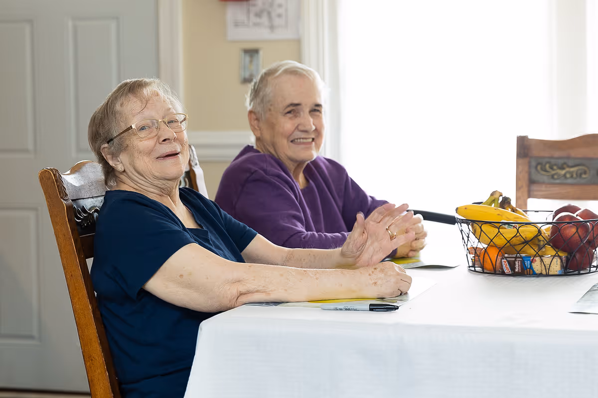 Two elderly women sitting at a dining table with a white tablecloth, smiling and looking towards the camera. On the table is a wire basket filled with bananas, apples, oranges, and other fruits. The room is well-lit with natural light coming from a window behind them.