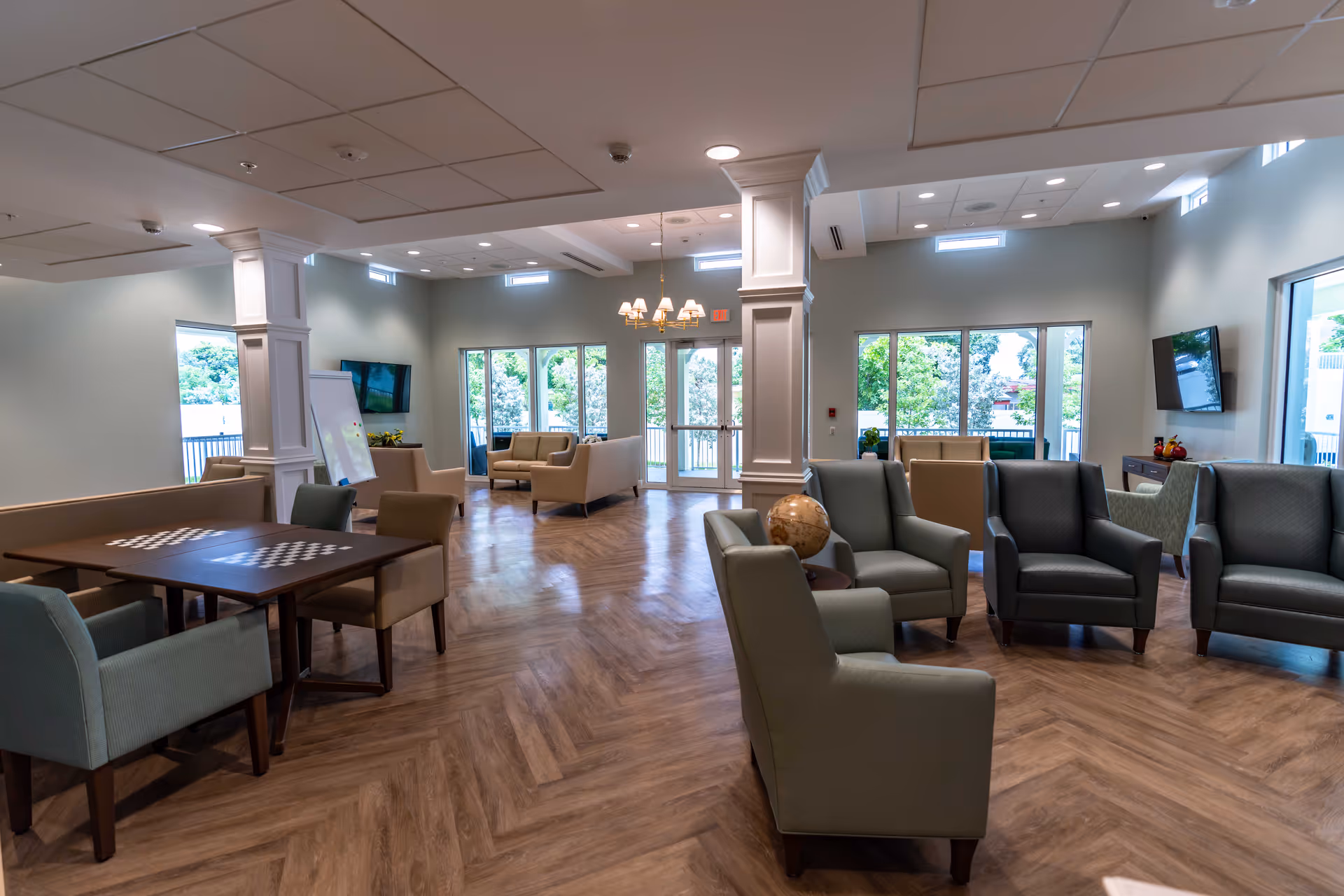 A spacious senior living common area with multiple seating arrangements including armchairs and sofas. There are large windows letting in natural light, a chandelier hanging from the ceiling, two wall-mounted TVs, and a table with chairs featuring checkerboard patterns. The room has wooden flooring and white pillars.