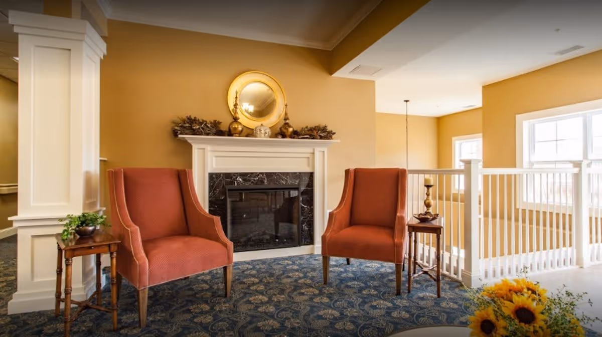 A cozy sitting area with two red upholstered armchairs placed on a patterned blue carpet in front of a white fireplace with a black marble surround. Above the fireplace is a round gold-framed mirror and decorative items. To the right, there is a white railing with windows letting in natural light, and a small wooden side table with a candle holder between the chairs.