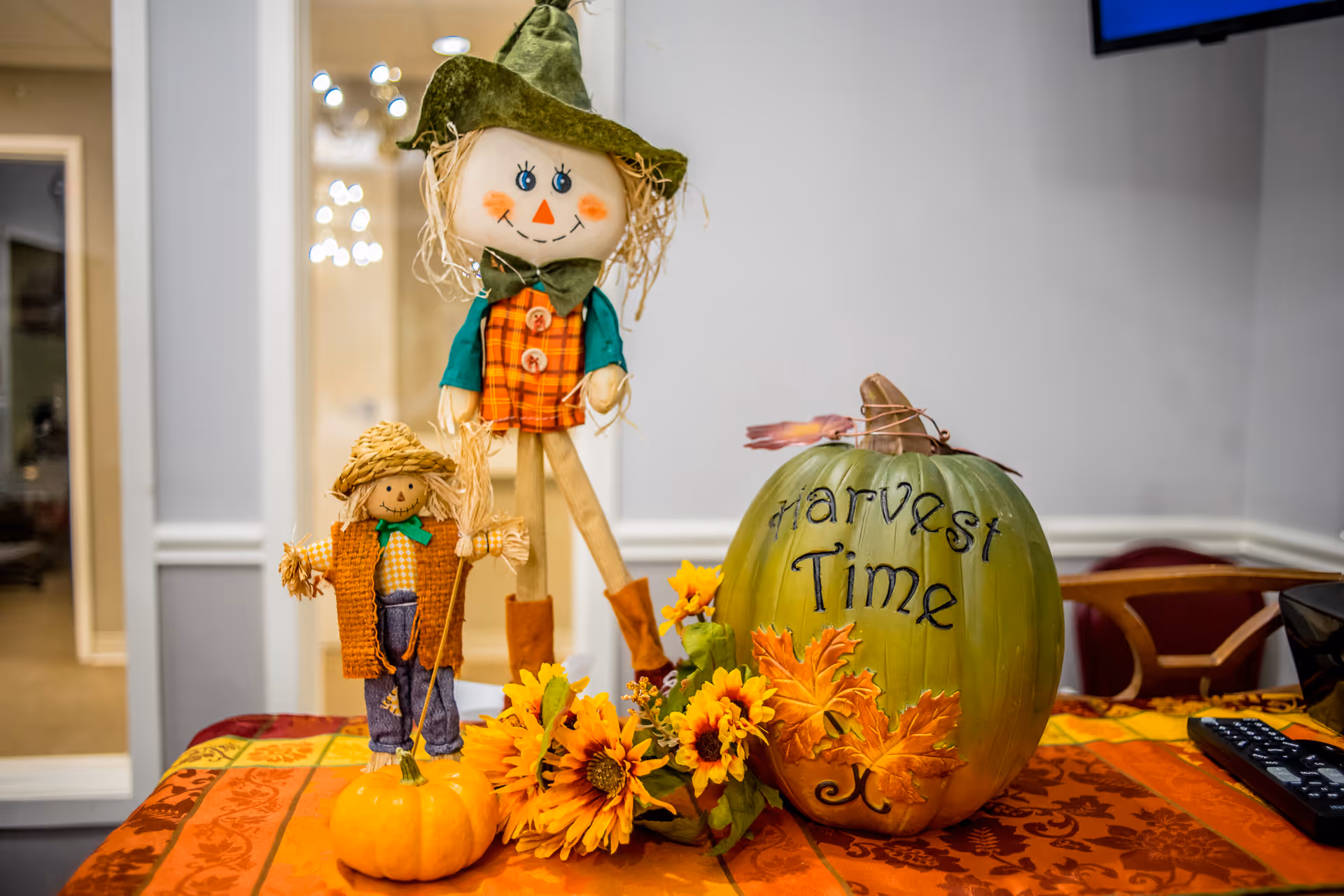 A festive autumn display on a table with an orange and red patterned tablecloth. The display includes two scarecrow dolls, one larger wearing a green hat and orange plaid outfit, and a smaller one with a straw hat and orange vest. There is a small orange pumpkin, a green pumpkin with the words 'Harvest Time' written on it, and artificial sunflowers and autumn leaves. A TV remote is visible on the right side of the table, and the background shows a light gray wall and part of a chair.