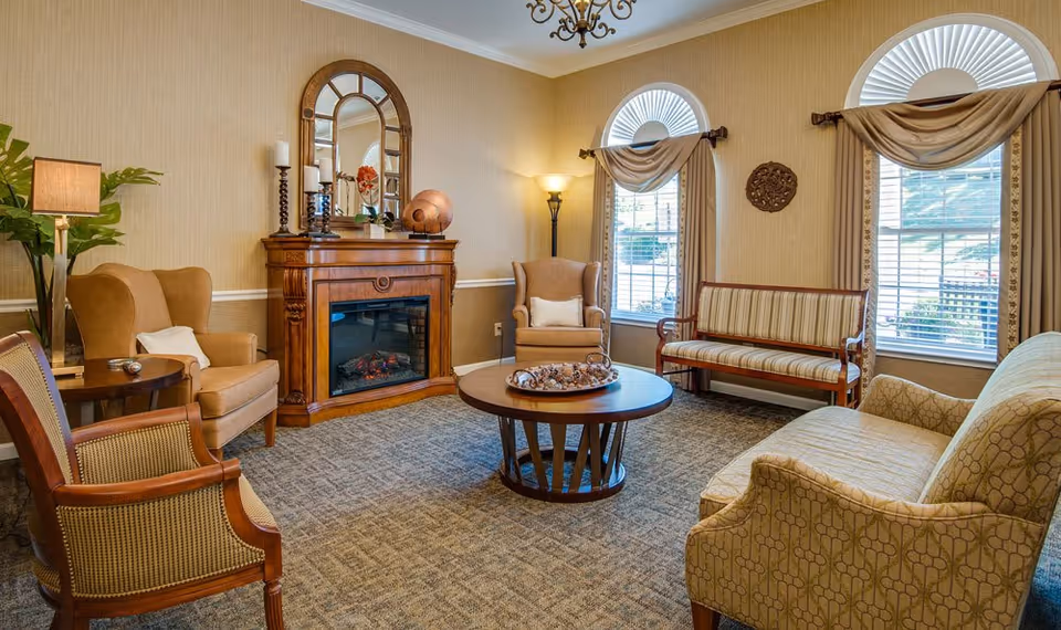 Cozy common living room with a fireplace, armchairs, sofa, round coffee table, and large arched windows with drapes.