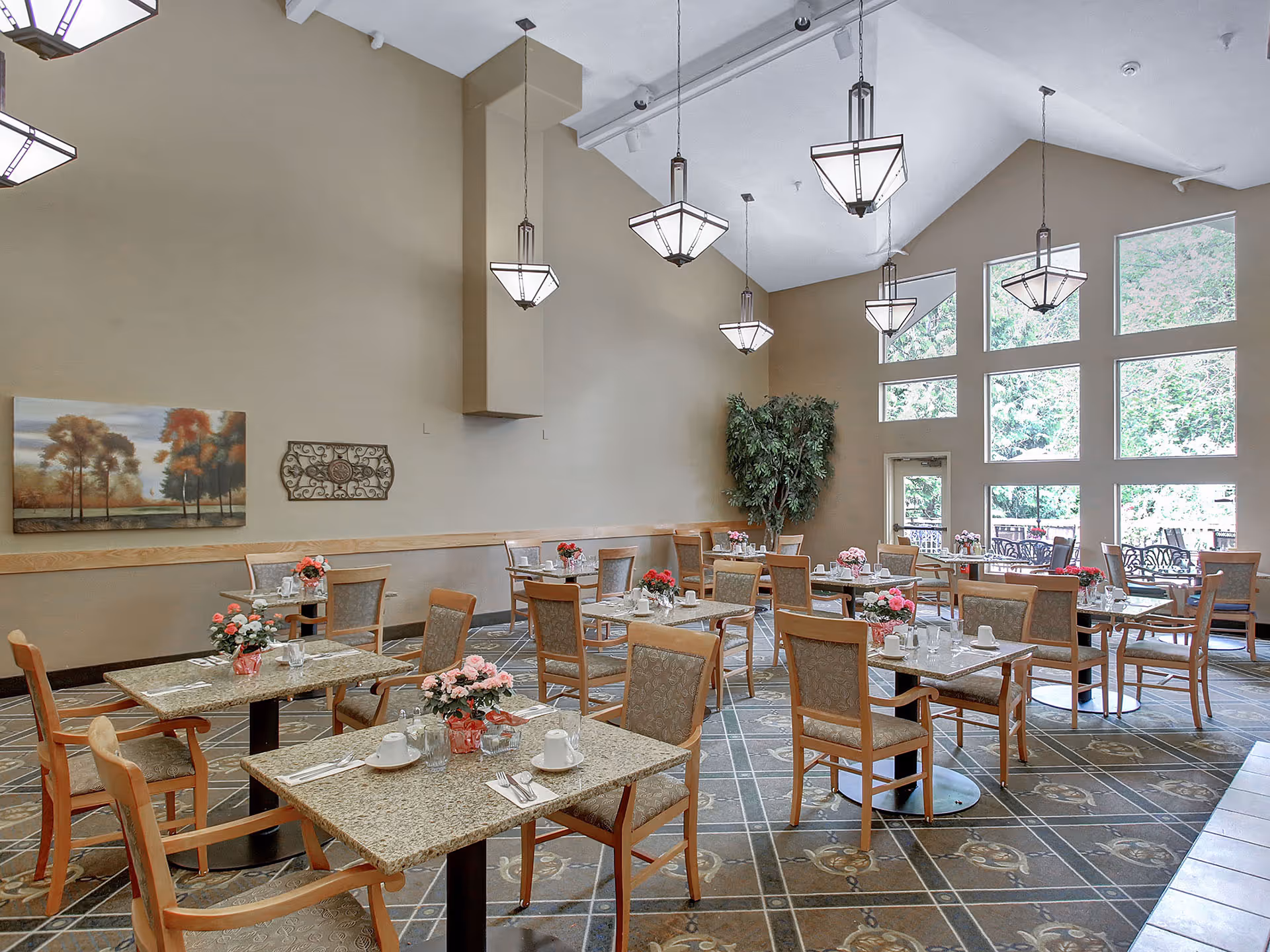 Spacious dining room with several tables set with cups and floral centerpieces under hanging lights and large windows.