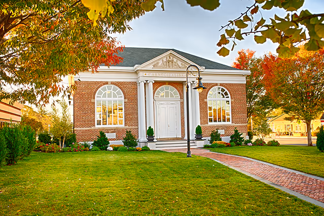 A brick building with white columns and large arched windows, surrounded by green grass and trees with autumn foliage. A brick pathway leads to the front door, and a vintage-style street lamp is positioned near the entrance.