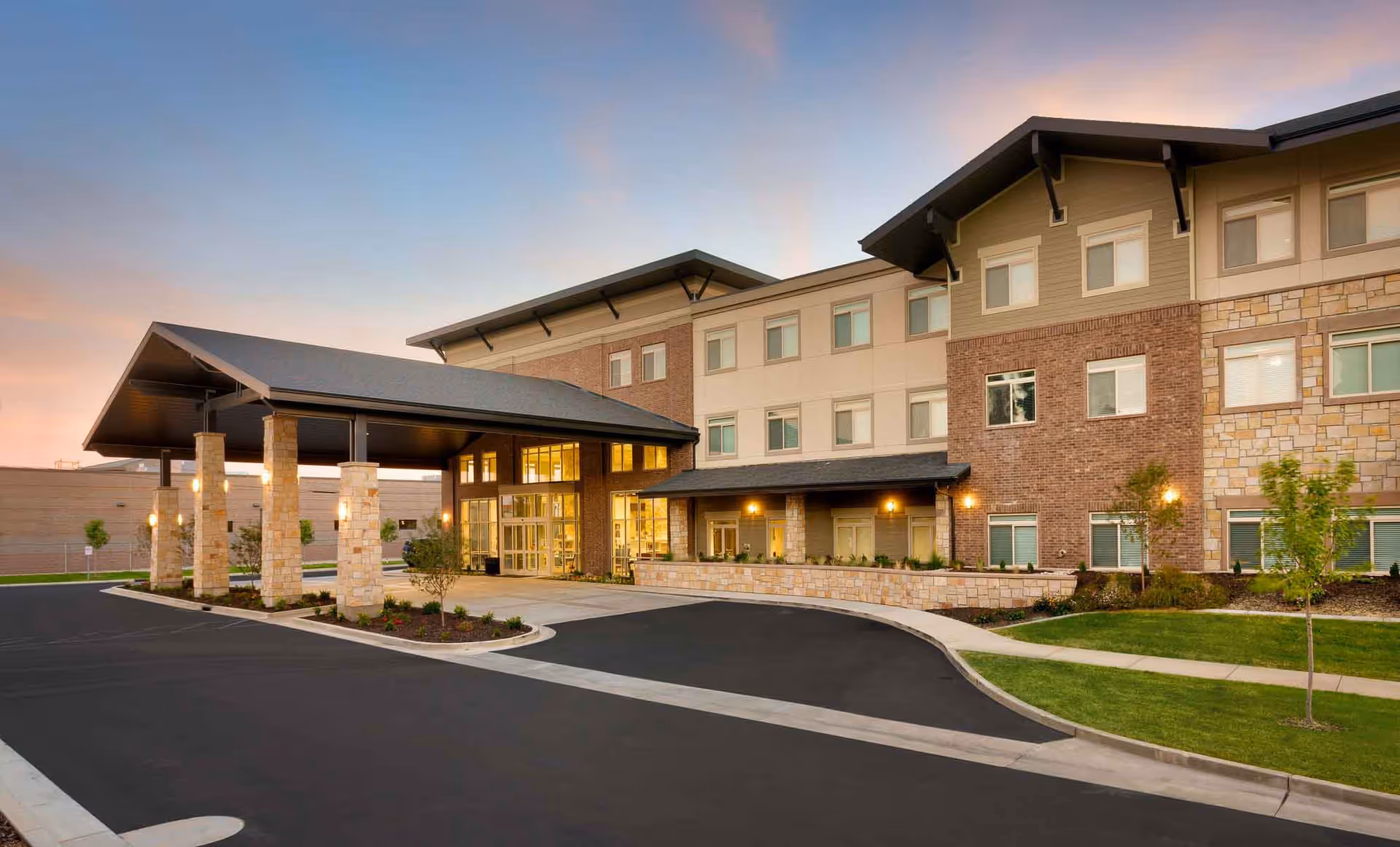 Exterior view of Creekside Assisted & Senior Living facility at dusk, showing a large three-story building with a covered entrance supported by stone pillars, well-maintained landscaping, and a paved driveway.