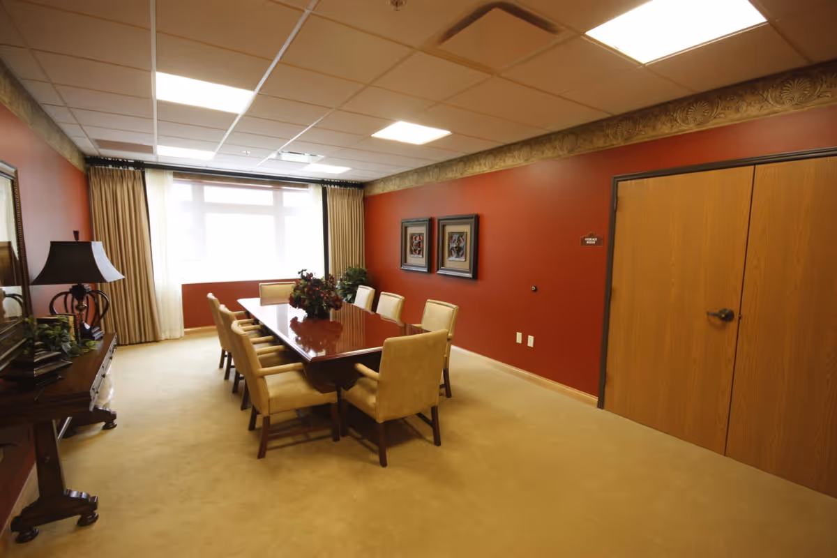 A conference or dining room with a long polished wooden table surrounded by eight beige upholstered chairs. The walls are painted a deep red with decorative molding near the ceiling. There are two framed artworks on the wall and a large window with beige curtains letting in natural light. A wooden side table with a lamp and some plants is on the left side of the room. A double wooden door is on the right wall with a small sign that reads 'STORAGE ROOM'.