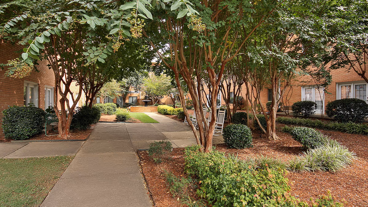 A landscaped outdoor courtyard area with a paved walkway, surrounded by trees, bushes, and plants. There are benches and rocking chairs along the sides, and brick buildings with windows in the background.