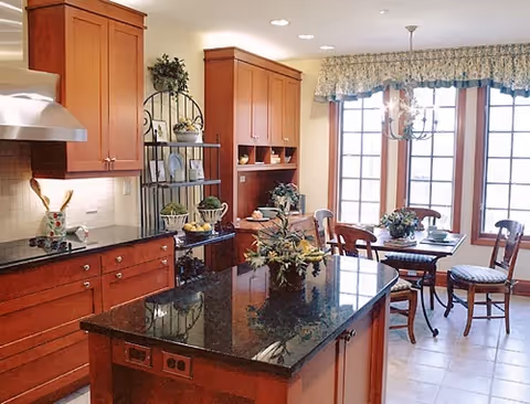 A bright kitchen with wooden cabinets, a black granite countertop island, and a dining area with a table and chairs near large windows with floral valances. The kitchen features a stainless steel range hood and decorative plants on the island and shelves.