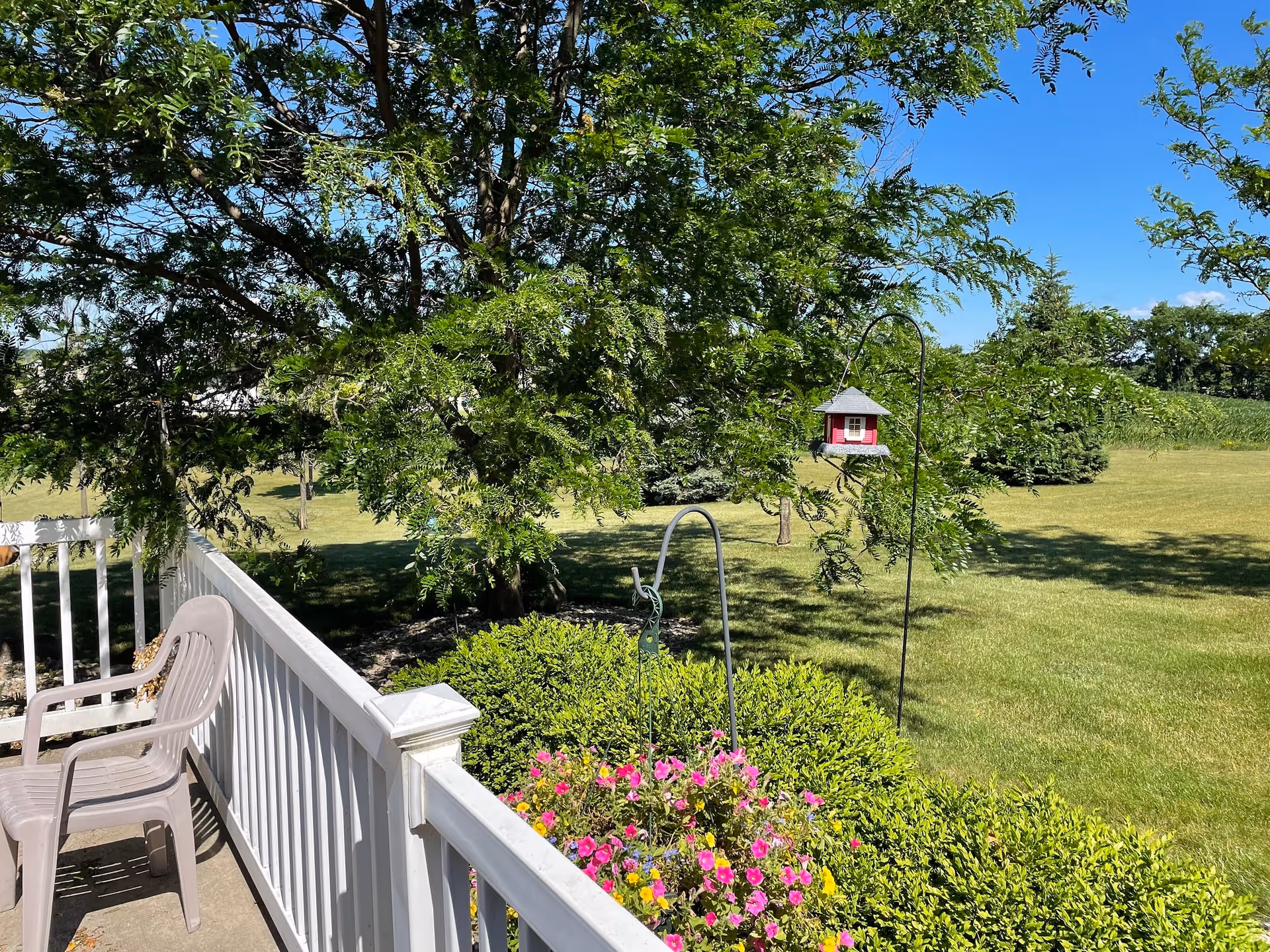 View from a white railing porch with a plastic chair, overlooking a green lawn with trees, bushes, a bird feeder, and colorful flowers under a clear blue sky.