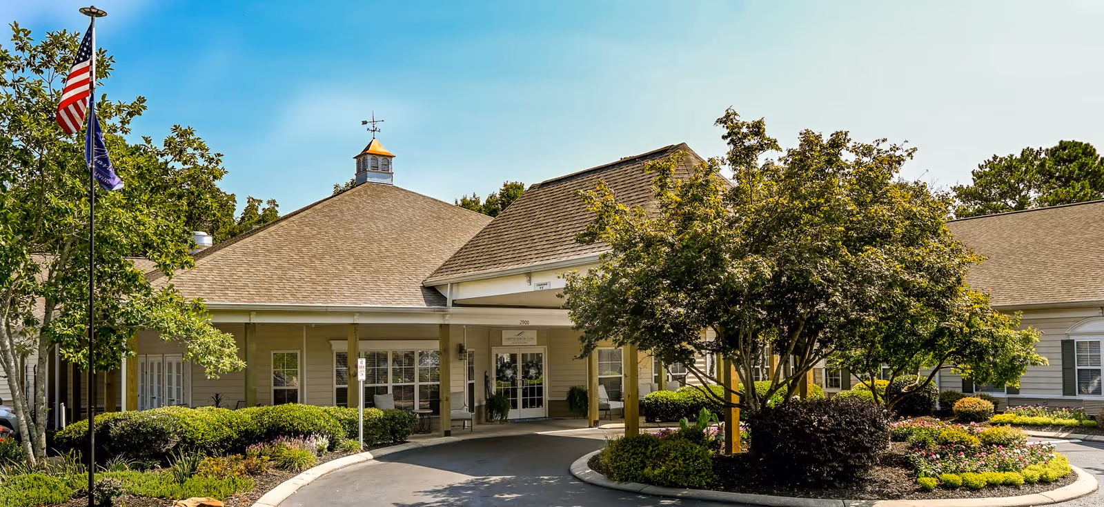 Exterior view of Charter Senior Living of Cleveland showing the entrance with a covered driveway, surrounded by landscaped greenery, trees, and flowers under a clear blue sky.
