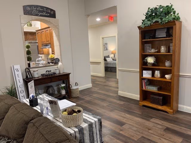 Interior view of a senior living facility showing a cozy common area with a brown couch, a wooden console table with decorative items, a tall wooden bookshelf with plants and decor, and a hallway leading to a bedroom. The flooring is wood, and the walls are painted light beige with white trim.