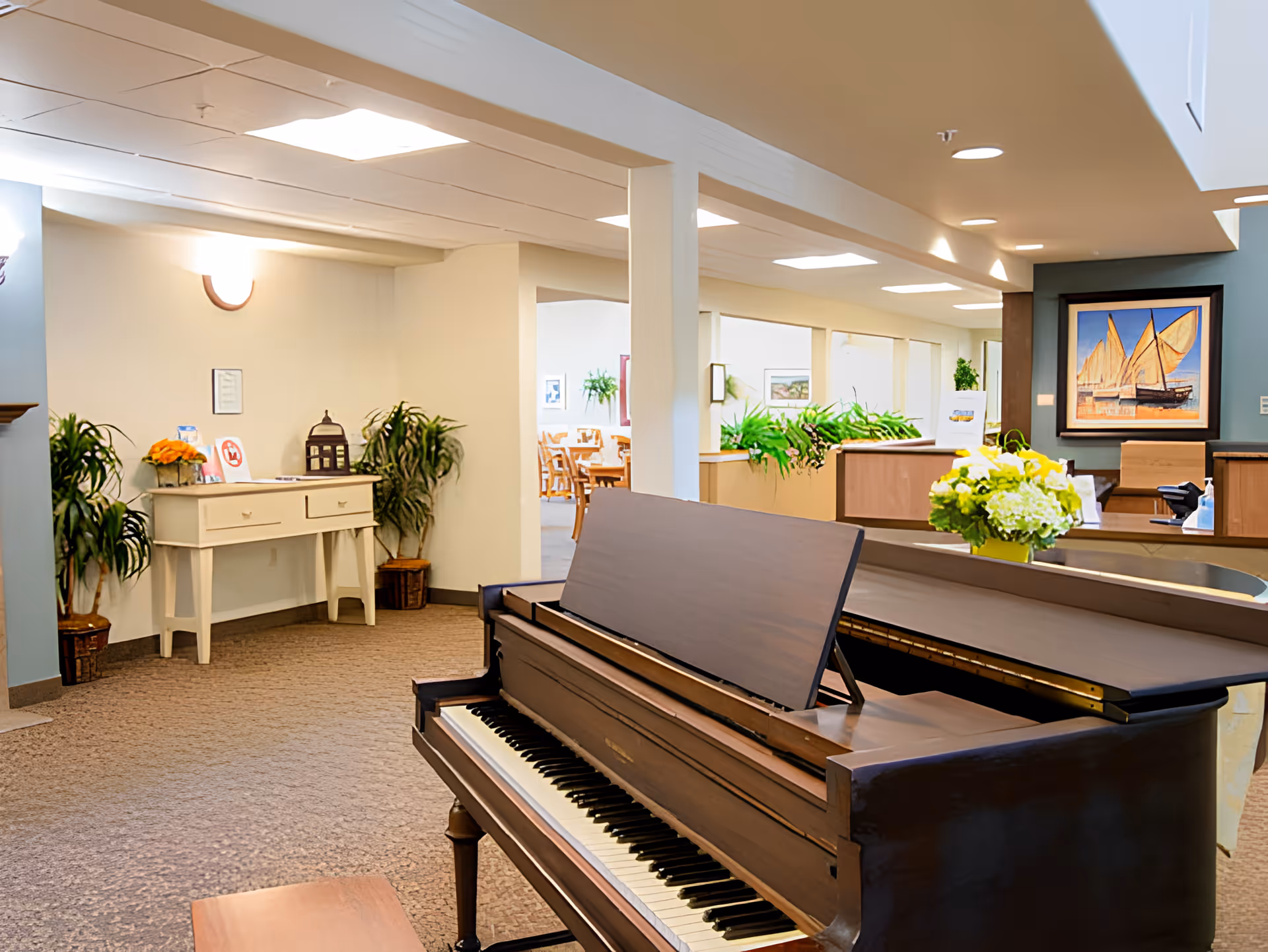 Interior view of a senior living facility featuring a grand piano in the foreground, a reception desk with a vase of flowers, potted plants, a small table with decorative items, and a painting of sailboats on the wall. The space is well-lit with ceiling lights and has a carpeted floor.