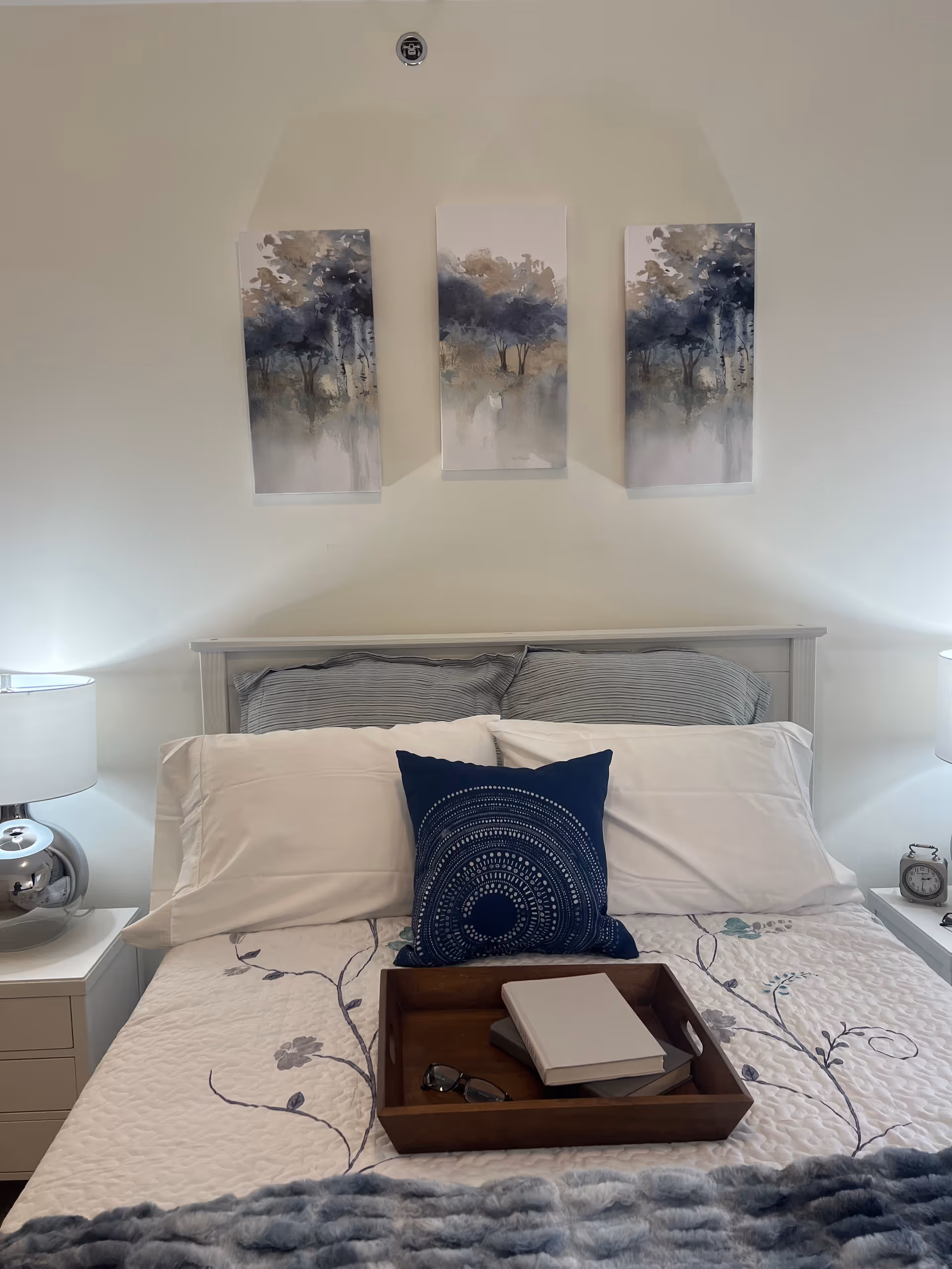A neatly made bed with white and gray pillows, a blue decorative pillow, and a floral-patterned quilt. On the bed is a wooden tray holding two books and a pair of glasses. Above the bed are three abstract landscape paintings with trees. There are two bedside tables with lamps and an alarm clock on the right table.