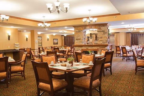 A spacious dining room in a senior living facility with multiple wooden tables and chairs arranged neatly. Each table is set with placemats, cups, and small plants. The room features warm lighting from ceiling chandeliers and wall sconces, a carpeted floor with a patterned design, and a stone fireplace in the center background.