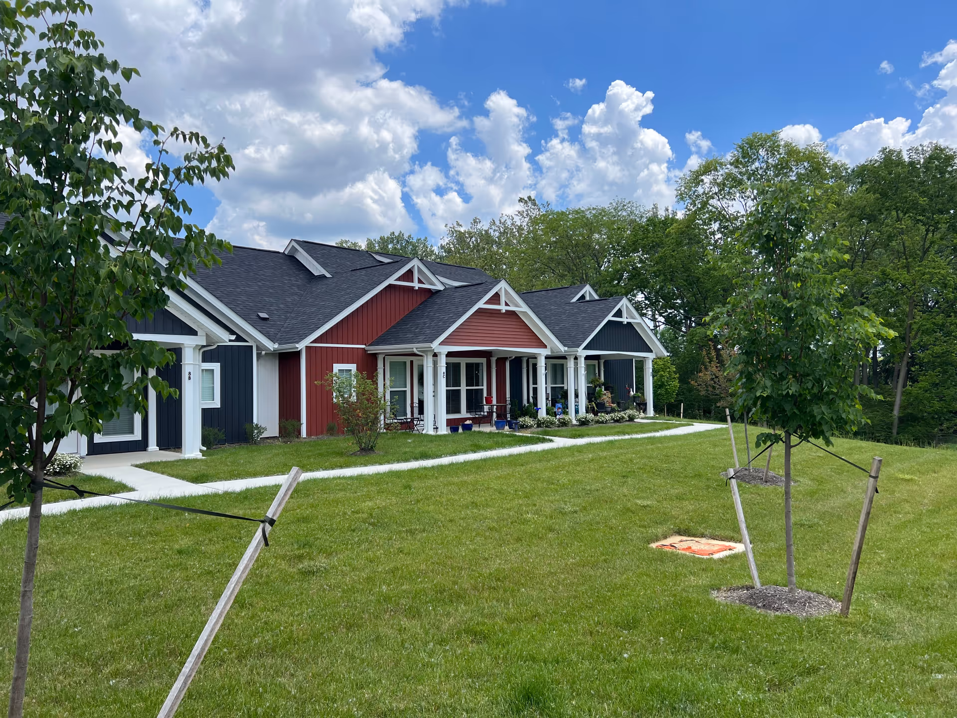 Front view of single-story red and gray cottage-style units with porches, a green lawn and young trees under a partly cloudy sky.